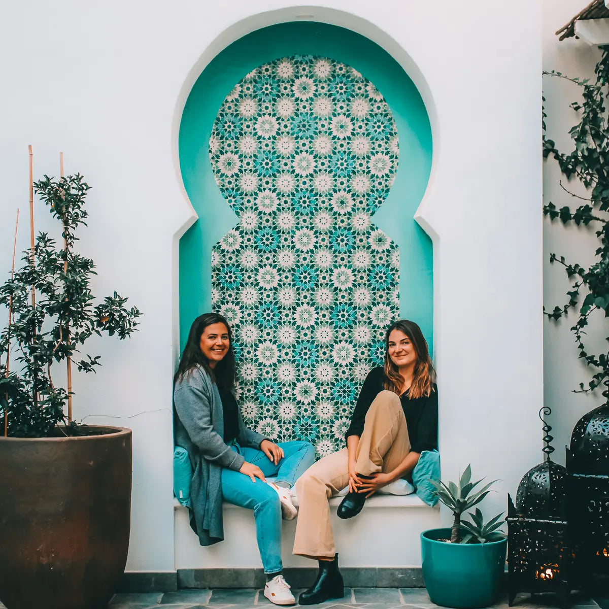 Zwei Frauen sitzen auf dem Boden vor einer dekorativen Wand mit türkisfarbenem Mosaik. Beide wirken entspannt und sind die Gründerinnen von Coffeebridge.